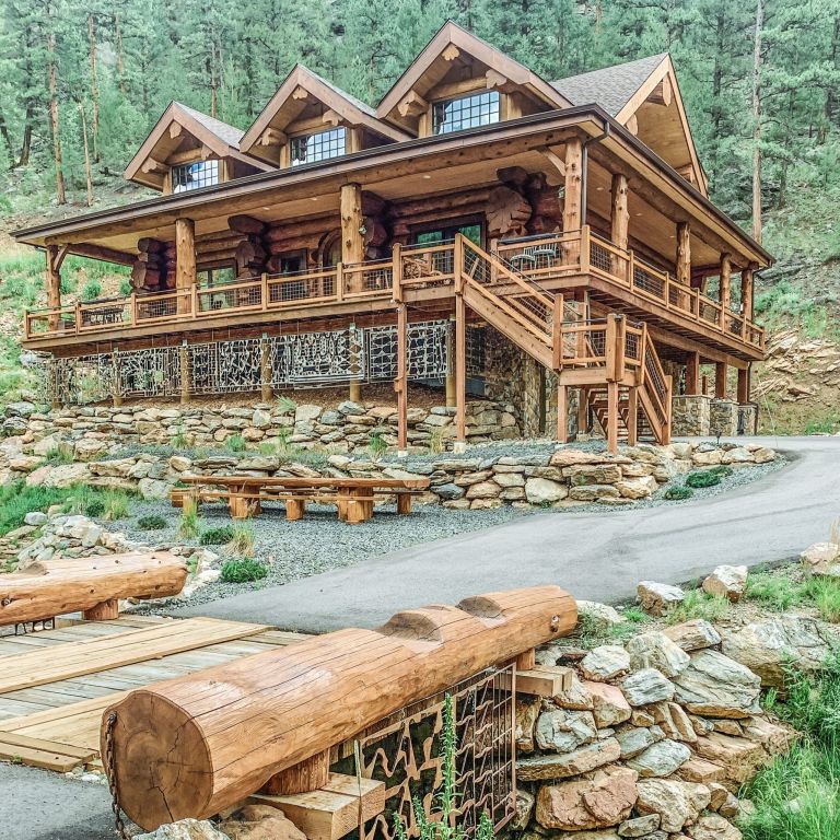 Western Red Cedar log home in Colorado with three dormers, full wraparound deck, stone base, and timber stair entry on a forested slope