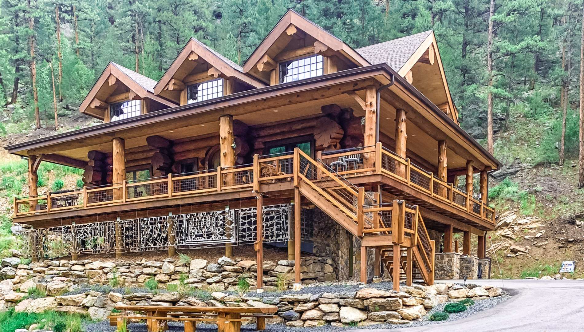 Western Red Cedar log home in Colorado with three dormers, full wraparound deck, stone base, and timber stair entry on a forested slope